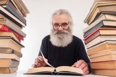 Weiser alter Mann mit grauen Haaren und Bart mit Brille, der ein Buch liest, in dem Bücher auf beiden Seiten gestapelt sind - © 123rf.com / grispb